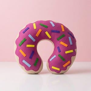 Op de afbeelding: Een reis kussen in de vorm van een donut. Het kussen heeft een magenta glazuur met kleurrijke rechthoekige hagelslag. De hagelslag is in tinten geel, groen, blauw en oranje. De basis van de donut is lichtbruin.