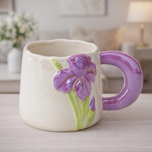 May include: Cream ceramic mug featuring a sculpted purple iris flower and green leaves. The handle is a matching shade of purple. The mug has a wide, rounded shape, ideal for tea or coffee. A decorative floral accent adds a touch of elegance to this kitchenware.