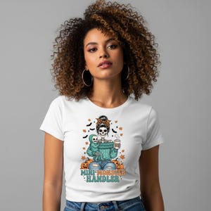 Puede incluir: Camiseta blanca con un diseño de Halloween. El diseño presenta un esqueleto sosteniendo un bebé esqueleto, rodeado de calabazas, murciélagos y hojas. El texto "MINI-MONSTER HANDLER" está impreso debajo.