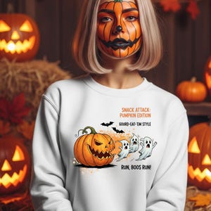 Puede incluir: Sudadera blanca con un diseño de Halloween. El diseño presenta una calabaza naranja, tres fantasmas de dibujos animados y murciélagos. El texto en la camiseta dice: "SNACK ATTACK: PUMPKIN EDITION, GOURD-EAT-'EM STYLE, RUN, BOOS RUN!"