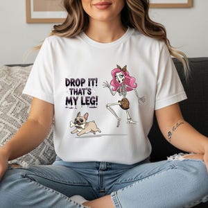 May include: White t-shirt with a graphic design featuring a skeleton with pink hair and a leopard print skirt, a running French bulldog with a bone, and the text "DROP IT! THAT'S MY LEG!"
