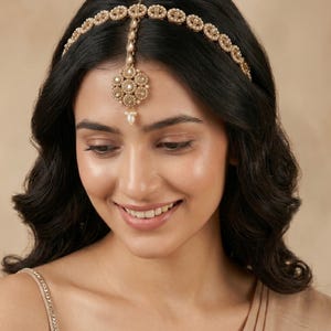 May include: A gold-toned headpiece featuring a delicate chain band adorned with small white pearls and circular filigree motifs. A central circular pendant with a floral design, inset with clear stones and a single dangling pearl, rests on the forehead.