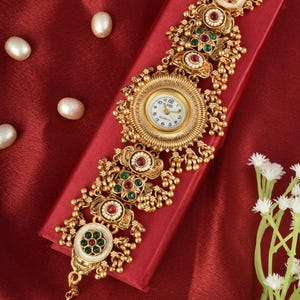 May include: A gold-toned quartz watch bracelet featuring a central round clock face with a white dial and black numerals. The band is composed of ornate metal links decorated with inlaid red and green stones, white accents, and small dangling beads.