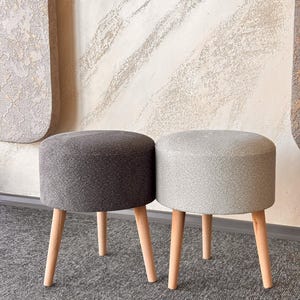 Puede incluir: Dos pequeños taburetes redondos tapizados. Uno es gris oscuro, el otro es gris claro. Ambos tienen patas de madera de color claro. Los taburetes están colocados uno al lado del otro sobre una alfombra gris, con una pared texturizada al fondo.