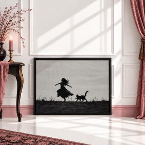 May include: A rectangular black-framed wall art print featuring a black and white silhouette of a person in a flowing dress running alongside a cat across a field. The scene is set against a light grey sky with sparse vegetation along the horizon.