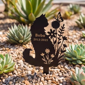 May include: A black metal garden stake featuring a silhouette of a cat sitting next to flowers and a butterfly. The cat's body is engraved with the name Bobo and the years 2013-2025. The design includes a pointed base for insertion into the ground.