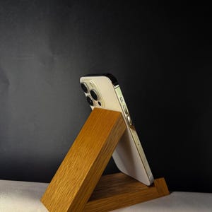 Puede incluir: Un soporte de teléfono de madera en forma triangular, sosteniendo un smartphone blanco. El soporte es de madera marrón claro con vetas visibles, sobre un fondo oscuro. El teléfono está inclinado para su visualización.