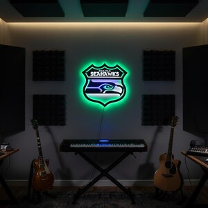 K&ouml;nnte beinhalten: Ein Neonschild mit dem Logo der Seattle Seahawks, einem Schild mit gr&uuml;nem Umriss und dem Teamnamen. Das Schild ist an einer grauen Wand in einem Musikstudio angebracht, mit Gitarren und einer Tastatur sichtbar.