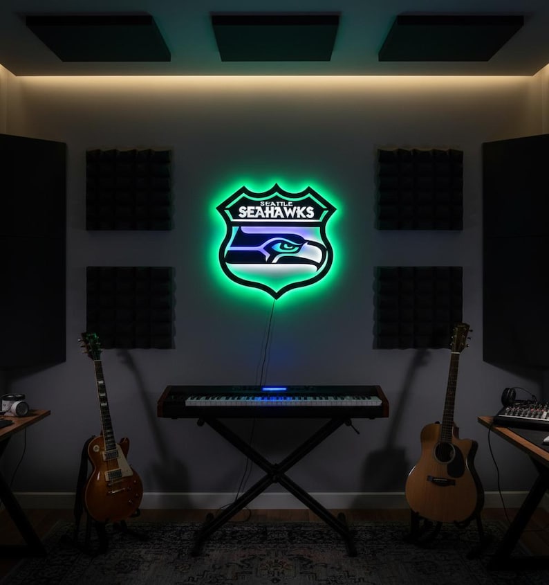 K&ouml;nnte beinhalten: Ein Neonschild mit dem Logo der Seattle Seahawks, einem Schild mit gr&uuml;nem Umriss und dem Teamnamen. Das Schild ist an einer grauen Wand in einem Musikstudio angebracht, mit Gitarren und einer Tastatur sichtbar.