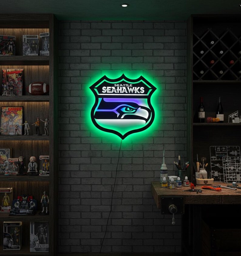 K&ouml;nnte beinhalten: Ein Neonschild mit dem Logo der Seattle Seahawks, einem stilisierten Adlerkopf in einem Schild. Das Schild hat einen schwarzen Umriss mit einem leuchtenden gr&uuml;nen Schein, und der Adler ist in Blau- und Wei&szlig;t&ouml;nen gehalten. An einer Backsteinmauer montiert.
