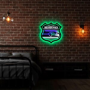 K&ouml;nnte beinhalten: Ein Neonschild mit dem Logo der Seattle Seahawks, einem Schild mit dem Teamnamen und einem stilisierten Falkenkopf. Das Schild hat eine gr&uuml;ne Umrandung und blaue und wei&szlig;e Akzente, montiert an einer Backsteinmauer.