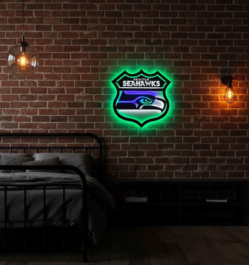 K&ouml;nnte beinhalten: Ein Neonschild mit dem Logo der Seattle Seahawks, einem Schild mit dem Teamnamen und einem stilisierten Falkenkopf. Das Schild hat eine gr&uuml;ne Umrandung und blaue und wei&szlig;e Akzente, montiert an einer Backsteinmauer.