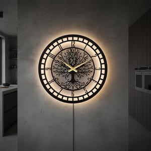 May include: A black, round wall clock with a tree of life design. The clock has Roman numerals and gold-colored hands. It is illuminated with a warm light, creating a halo effect around the clock face. The clock is mounted on a gray wall.