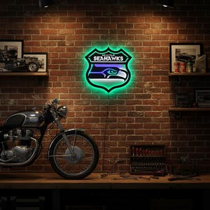 K&ouml;nnte beinhalten: Ein Neonschild mit dem Logo der Seattle Seahawks, einem Schild mit einem stilisierten Adlerkopf in Blau, Gr&uuml;n und Wei&szlig;. Das Schild ist an einer Backsteinmauer angebracht, mit einem Motorrad und Werkzeugen im Vordergrund. Das Schild leuchtet.
