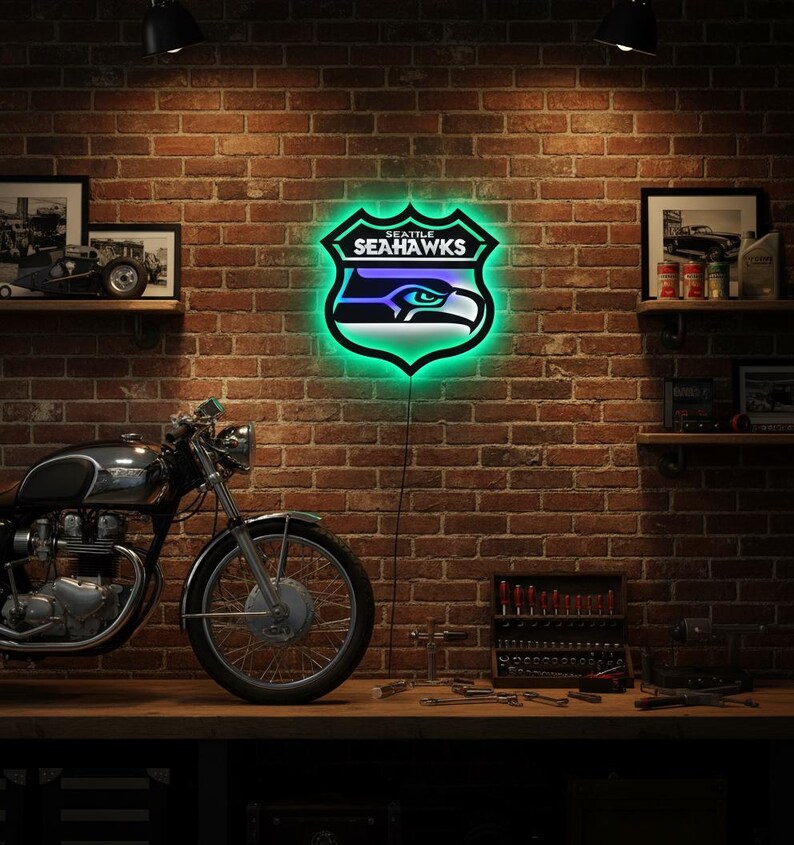 K&ouml;nnte beinhalten: Ein Neonschild mit dem Logo der Seattle Seahawks, einem Schild mit einem stilisierten Adlerkopf in Blau, Gr&uuml;n und Wei&szlig;. Das Schild ist an einer Backsteinmauer angebracht, mit einem Motorrad und Werkzeugen im Vordergrund. Das Schild leuchtet.