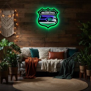 K&ouml;nnte beinhalten: Ein Neonschild in Form des Logos der Seattle Seahawks, mit gr&uuml;nem Umriss und dem Teamnamen. Das Schild ist an einer Holzwand &uuml;ber einem Sofa mit Kissen und Decken angebracht. Der Raum ist mit Pflanzen und einem Jute-Teppich dekoriert.