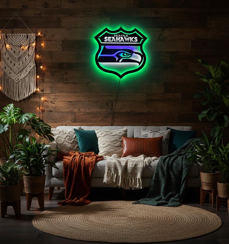 K&ouml;nnte beinhalten: Ein Neonschild in Form des Logos der Seattle Seahawks, mit gr&uuml;nem Umriss und dem Teamnamen. Das Schild ist an einer Holzwand &uuml;ber einem Sofa mit Kissen und Decken angebracht. Der Raum ist mit Pflanzen und einem Jute-Teppich dekoriert.