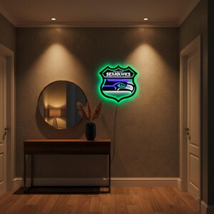 K&ouml;nnte beinhalten: Ein Neonschild in Form eines Schildes mit dem Logo der Seattle Seahawks. Das Schild hat eine gr&uuml;ne Umrandung und leuchtet in den Farben des Teams. Es ist an einer Wand in einem Flur mit Holzboden und einem dunklen Holztisch angebracht.