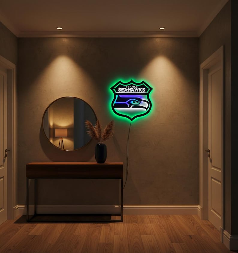 K&ouml;nnte beinhalten: Ein Neonschild in Form eines Schildes mit dem Logo der Seattle Seahawks. Das Schild hat eine gr&uuml;ne Umrandung und leuchtet in den Farben des Teams. Es ist an einer Wand in einem Flur mit Holzboden und einem dunklen Holztisch angebracht.