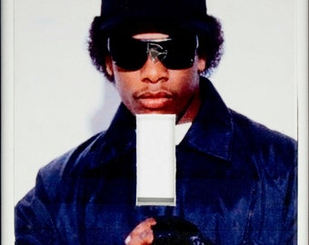 May include: A black and white photograph of a man wearing a black baseball cap with the word "Compton" written in white letters. He is wearing sunglasses and a black jacket. The photo is printed on a white light switch cover.