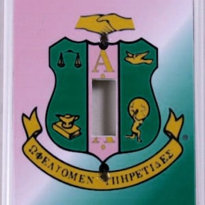 May include: A light switch cover with a green and pink background featuring a crest with a gold border and the Greek letters "ΦΕΑΤΩΜΕΝ ΙΗΡΕΤΙΣΔΕΣ".