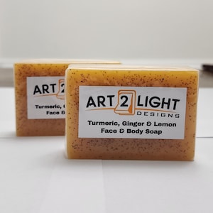 May include: Two rectangular bars of orange-toned soap with visible dark brown specks. Each bar features a white label with the text ART 2 LIGHT DESIGNS and Turmeric, Ginger & Lemon Face & Body Soap printed in black ink on the front surface.