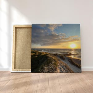 Puede incluir: Impresión en lienzo de un paisaje costero con puesta de sol. La obra muestra una playa arenosa, dunas cubiertas de hierba y un cielo vibrante con nubes. El lienzo se exhibe junto a su marco de madera.