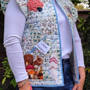 May include: A quilted vest with a floral pattern in light blue, white, and green. The vest features a squirrel and mushroom applique, a crocheted feather, and a pink flower. The text "I have pockets!" is visible.