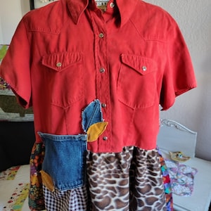 May include: A red short-sleeved shirt with two chest pockets and a button-down front. The shirt is paired with a patchwork skirt featuring denim, leaf appliqués, a brown and white animal print, and a black and white checkered pattern.
