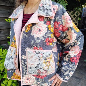 May include: A quilted jacket featuring a dark gray base with a vibrant floral pattern in shades of pink, red, and yellow. The garment includes a light pink trim, a wide collar, and two large front pockets with a white snakeskin-like texture.