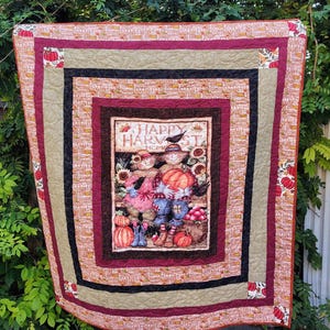 Op de afbeelding: Een herfstquilt met een centraal paneel met vogelverschrikkers die pompoenen en zonnebloemen vasthouden, met de tekst "Happy Harvest Home". De quilt heeft een gelaagd ontwerp met bordeauxrode, zwarte en bruine randen en is versierd met pompoenmotieven.
