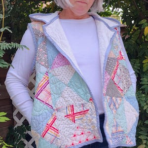 May include: A quilted vest featuring a patchwork design in pastel colors, including blue, pink, and white. The vest has a light blue trim and a white interior. The person is wearing a white long-sleeve shirt and blue jeans.