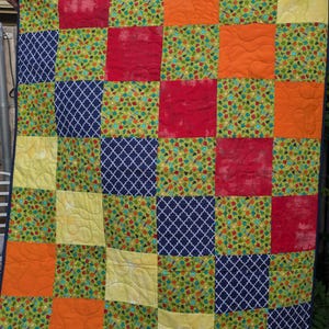May include: A patchwork quilt with a variety of colorful squares. The quilt features squares of red, orange, yellow, navy blue, and green fabric. Some squares have a floral pattern, while others have a geometric design. The quilt is held up by two hands.