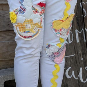 May include: White denim jeans with colorful patchwork designs, including a heart, a flower, and a geometric pattern. The jeans have a yellow zig-zag trim down the side.