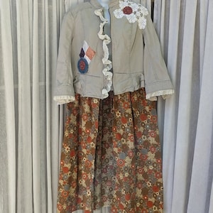 May include: A beige jacket with a crocheted lace trim and a patchwork design on the front. The jacket is worn over a brown floral skirt.