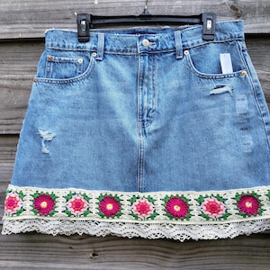 May include: A blue denim skirt featuring a distressed finish, a button and zipper closure, and five pockets. The hem is decorated with a wide band of colorful floral crochet squares in pink, red, and green, finished with a white lace trim.