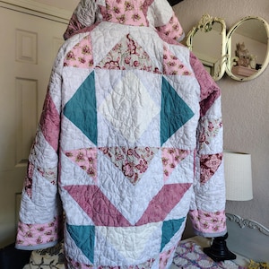 May include: A quilted jacket with a hood, featuring a patchwork design in white, pink, and teal. The jacket has a floral pattern on the hood and some patches. The overall design is a mix of geometric shapes and floral prints.