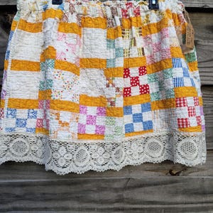 May include: A skirt crafted from a patchwork quilt featuring a grid of yellow, white, red, blue, and pink patterned fabric squares. The garment has an elasticized waistband and a wide, intricate white lace trim along the bottom hemline.