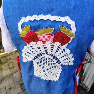 May include: A blue vest featuring a central applique of a white crocheted basket filled with three fabric strawberries. The strawberries have green fabric leaves and red bodies, with one featuring a red and white gingham pattern. Red ribbons tie at the sides.