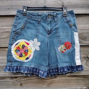 May include: Blue denim shorts with decorative patches and lace trim. The shorts feature a patchwork design on the front, with a ruffled hem and white lace accents. The shorts are hanging on a black hanger.