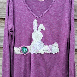 May include: A long-sleeved purple V-neck shirt with a decorative white bunny applique. The bunny is made of quilted fabric with floral and lace accents. The shirt is hanging on a wooden background.