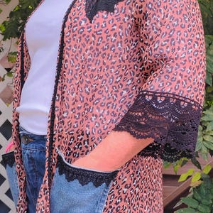 May include: A peach-colored leopard print kimono with black lace trim on the sleeves and pockets. The kimono features a black feather and blue denim accents on the shoulders. The pockets are made of denim.