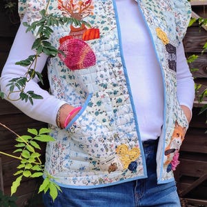 May include: A quilted vest with a floral pattern in light blue, white, and green. The vest features a blue trim and various appliqued patches, including a deer, a fox, and a yellow heart. The vest has a high collar and ruffled shoulders.