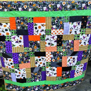May include: A patchwork quilt with a Halloween theme. The quilt features squares of fabric with various patterns, including gnomes, pumpkins, and stripes in orange, purple, black, and green. The quilt is bordered with a green fabric.