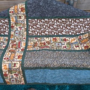 Peut inclure: Une courtepointe faite à la main sur le thème du camping. La courtepointe présente divers panneaux avec des images d'ours, de tentes et d'équipement de camping. La palette de couleurs comprend le marron, le vert et le bleu, avec un motif en forme de pierre au centre.