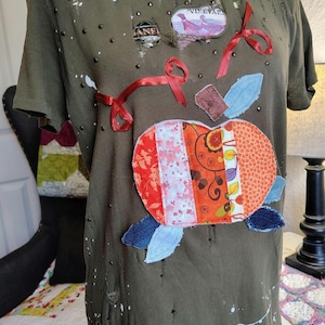 May include: Olive green t-shirt with a patchwork design. The shirt features a large, colorful pumpkin applique with red ribbon accents and denim leaf details. The shirt has distressed details and small embellishments.