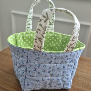 May include: A small, quilted fabric basket with two handles. The exterior is light blue with a floral pattern, while the interior is green with white polka dots. The handles are white with a leaf design.