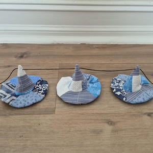 May include: Three small, handmade witch hats with blue and white patchwork designs are strung together on a black string. The hats have a conical shape and are set against a wooden floor and a white wall.