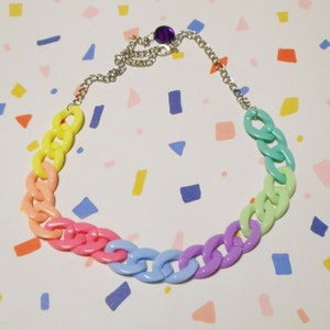 Peut inclure: Un collier en chaîne de plastique arc-en-ciel avec un fermoir argenté. Le collier est composé de maillons épais et comporte une perle violette au fermoir.