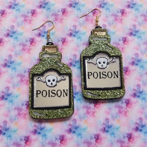 Peut inclure: Une paire de boucles d'oreilles pendantes en forme de flacons de potion avec une finition &agrave; paillettes vertes. Chaque flacon pr&eacute;sente une &eacute;tiquette argent&eacute;e avec une ic&ocirc;ne de cr&acirc;ne et d'os crois&eacute;s au-dessus du mot POISON en lettres noires.